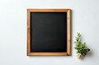 © Yuliia - Empty square chalkboard with wooden frame on white wall background, small green plant in corner for minimalist home decor.
