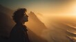 © Yuliia - Silhouette of young man with curly hair gazing at dramatic coastal mountain landscape during golden hour sunset with fog rolling over ocean waves.