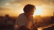 © swillklitch - Young Latino man with messy hair sits alone on city rooftop at sunset, thinking peacefully and enjoying the golden hour view in urban summer evening