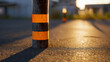 © Sergej Gerasimov - A black and orange striped bollard casting a long shadow on a textured asphalt road illuminated by warm sunset light with blurred background elements and urban detai