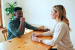 © Mediaphotos - Young black man sitting at wooden table facing young woman holding glass, both engaging in serious business conversation in modern office setting with plant in background