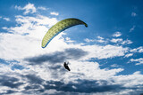 Paraglider soaring toward bright sunlight across a vast blue sky dotted with white and dark clouds, silhouette pilot enjoying high altitude freedom and summer adventure