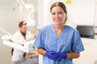 © JackF - Smiling female dentist assistant stands in a blue gown and gloves against the background of the office and a dental chair. Nurse invites clients to visit the dentist