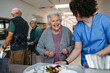 © Halfpoint - Senior woman having lunch with supportive caregiver in community center cafeteria.