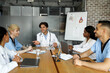 © Prostock-studio - Handsome man doctor with digital tablet talking to his colleagues, international group of positive doctors friendly professional team sitting at table, having morning meeting or brain-storming