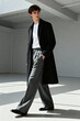 © cmapuk_0nline - A young stylish man in a black overcoat, white t-shirt, and wide grey trousers walks in a minimalist interior. Modern urban male fashion look.