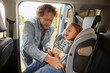 © Stockphotodirectors - A father joyfully assists his child in securing the seatbelt in a car seat, ensuring safety before heading out for a family adventure on a sunny day.