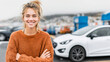 © Bonsales - Young woman smiling proudly outdoors, standing with a white car in the background, representing new car ownership and independence