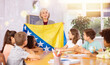 © JackF - Mature female teacher tells pupils the history of Bosnia and Herzegovina in class and holds the national flag of the country