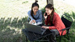 © NAMPIX - Two Asian women sit outdoors in a sunlit garden; one smiles while on a mobile phone call, looking at a laptop the other woman is pointing at.