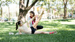 © NAMPIX - Two cheerful young Asian women exercising together in a park, with one performing a leg lift exercise on a mat while being encouraged by her friend.