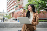 Pretty asian woman office worker holding coffee cup over laptop sitting crossed leg on bench in park