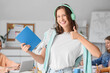 © Pixel-Shot - Female student with headphones and notebook showing thumb-up in classroom