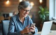 © Thomas - Senior mature experienced woman at workplace inside office, business woman happy smiling using phone sitting with laptop, gray haired woman boss using app on smartphone typing message. High quality