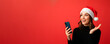 © Iuliia - Happy woman in Santa hat looking with smile at mobile phone in Christmas holiday. Communication on smartphone in holiday in studio on red background, banner