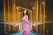 © BullRun - Smiling Hispanic woman in floral pink dress holding smartphone outdoors among golden art installation, symbolizing individuality, freedom, and digital connection in modern lifestyle.