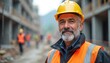 © miss irine - Construction worker with grey beard wears yellow hard hat and orange vest. He stands on building site. Other workers visible in distance on construction project.