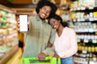 © Prostock-studio - A man and woman smile while shopping in a grocery store. The man holds a smartphone in front of them, displaying a blank screen. They look relaxed and happy together.