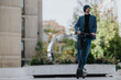 © qunica.com - A stylish man rides an electric scooter through a modern urban plaza. He wears a blue blazer, black turtleneck, and beanie, exuding confident, relaxed street style suitable for city living.