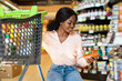 © Prostock-studio - A woman reviews a jar of sauce while sitting on a grocery cart in a store aisle. She enjoys exploring various products and is focused on her choice.
