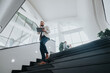 © qunica.com - A confident businessperson navigates a sleek, sunlit atrium, using a tablet while on a call. In the background, people chat near large windows, suggesting collaboration and a dynamic work environment.