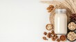 © lizahanifah - A glass jar filled with creamy plant-based milk sits on a woven mat, surrounded by ingredients like oats, walnuts, pecans, and wheat stalks. Natural light illum