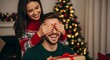 © Ali Raza - A Christmas surprise Woman covering man s eyes giving gift near decorated tree Holiday joy and romance captured in cozy