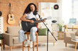 © Pixel-Shot - Pretty young African-American woman with microphone sitting and playing acoustic guitar in living room