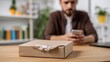 © imagemir - A young man of Middle-Eastern descent examines a damaged package on a wooden table while using his smartphone.