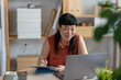 © lordn - Smiling Asian woman entrepreneur writing notes while holding a coffee mug, working at her laptop in a calm and organized home office