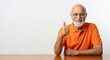 © PRASANNAPIX - A cheerful elderly Indian man with a white beard and glasses gives a thumbs-up while smiling, seated at a table in an orange shirt.