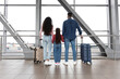 © Prostock-studio - A family of three stands at an airport window, gazing outside at the runway. The mother and father hold suitcases while their daughter admires the view.