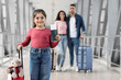 © Prostock-studio - A young girl stands in an airport terminal holding a passport and a teddy bear. Behind her, a woman and man are getting ready for their trip, each carrying luggage.