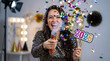 © PhotoGranary - Happy woman celebrating New Year 2026 with a confetti cannon and a colorful number prop. Festive party scene featuring a young female in a sequin dress having fun in a photo booth setting.