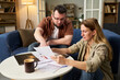 © pressmaster - Caucasian middle aged man and Caucasian middle aged woman reviewing tax documents together at table, man pointing at paper while woman holding paperwork, laptop and calculator visible
