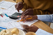 © pressmaster - Closeup of Black man and Black woman calculating taxes together using calculator and reviewing financial documents at table, hands holding pencil and receipts, paperwork scattered around