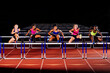 © Connect Images - Five women competing in a hurdles race on a track at night. WA, USA