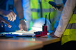 © ultramansk - A close up of an engineering team collaborating at night. Hands point at a glowing blueprint on a light table, with a hardhat and walkie talkie on the desk.