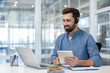© Liubomir - Happy bearded man wearing a headset and working remotely from an office, providing online customer support and technical assistance while looking at a laptop screen and holding a digital tablet