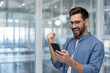 © Liubomir - Young man in blue shirt standing in a modern office, expressing joy and excitement while looking at his smartphone, celebrating a significant achievement or good news