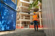 © DusanJelicic - Construction worker wearing safety vest and helmet walking on construction site carrying toolbox and tools, smiling towards camera with scaffolding and temporary buildings in background
