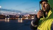© ViskBx - Man in safety vest holding a walkie talkie at dusk with trucks in the background at a shipping yard