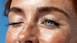 © Yuliia - Close-up of young woman with freckles winking against blue background, showing one bright blue eye and natural lashes.