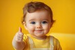 © btiger - Happy baby with blue eyes giving thumbs up wearing yellow clothes against a bright yellow background