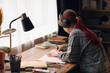 © DragonImages - Caucasian teenage girl with long red hair wearing headphones sitting at desk studying and writing on paper surrounded by school supplies in bedroom with sunlight coming through window