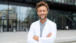 © Nattapol - Confident handsome male doctor smiling outside modern hospital. professional man in white coat with arms crossed looking at camera