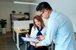 © Mediaphotos - Young adult Asian woman sitting at desk reviewing documents with young adult Asian man standing beside her holding clipboard and pen, both collaborating in modern office workspace