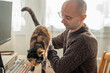 © kittyfly - bald man in brown sweater gently holding tortoiseshell cat on desk, working computer with keyboard and monitor, in cozy home office setting, petting cat during work break, furry companion nearby