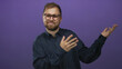 © Krakenimages.com - Bearded young man with glasses presenting open hands palm up gesture in studio with purple backdrop and navy shirt; curious thought.