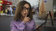 © Krakenimages.com - Transgender woman wearing glasses holds tablet with finger to lips on busy city street; secretive hush.